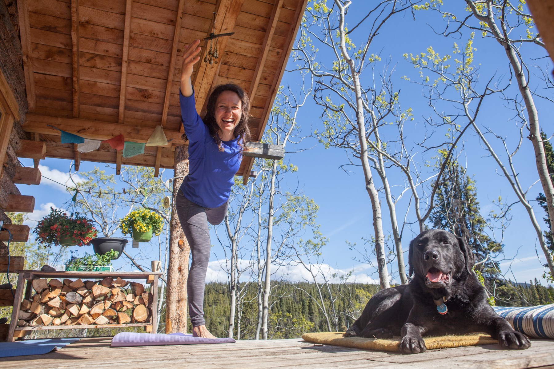 Someone doing a yoga pose on the porch of a cabin with a cute black lab looking on.